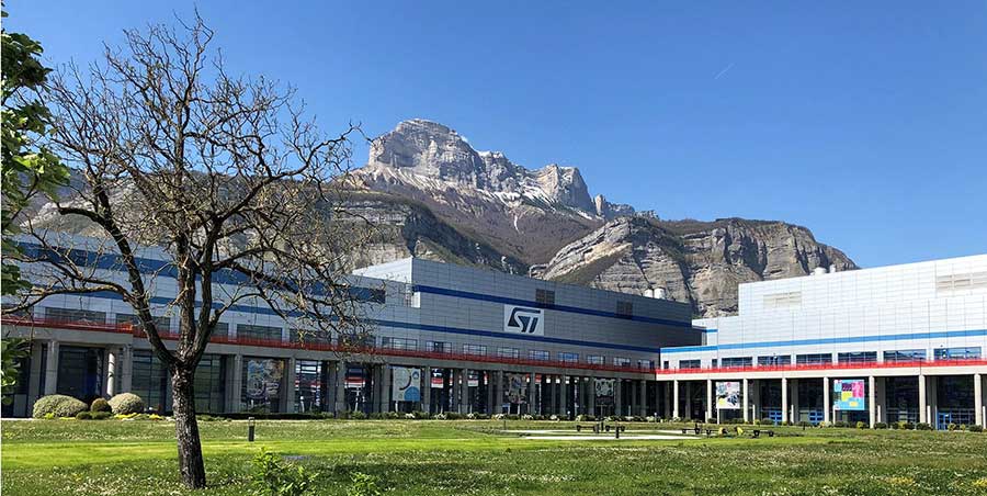 A picture of STMicroelectronics' chip fab in Crolles, France, with two gray buildings sitting below a mountain.